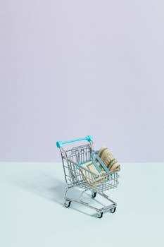 A small shopping cart holding rolled dollar bills on a pastel background, symbolizing online shopping trends.