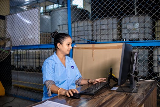 A woman in blue uniform operates a computer inside a factory warehouse, focusing on her task.