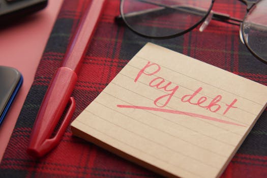 Close-up of a note reading 'Pay debt' next to a red pen on a plaid fabric, emphasizing financial reminders.