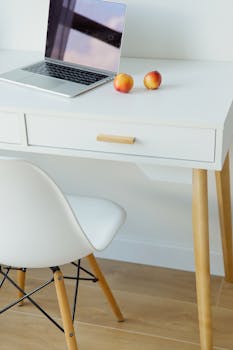 Modern minimalist workspace featuring a sleek laptop and two fresh apples on a white desk with a white chair.