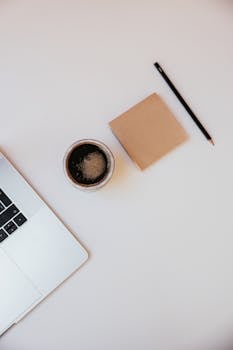 Stylish overhead view of a laptop, coffee cup, notepad, and pencil on a white desk.